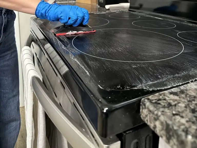 Gloved hand scrubbing a glass cooktop with a scraper pad during a house cleaning